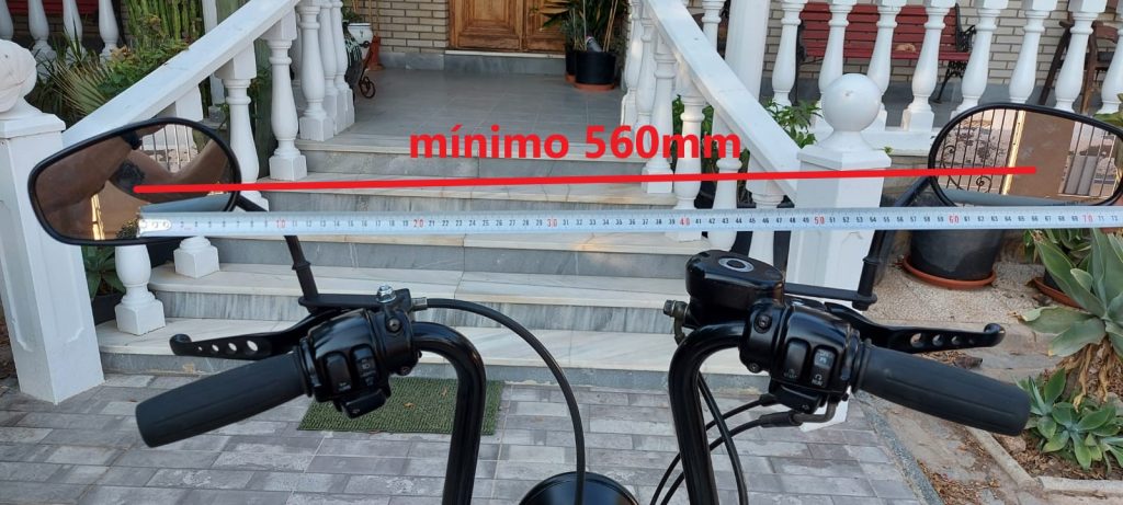 Imagen frontal tomada desde la posición del conductor de una motocicleta, mostrando los retrovisores exteriores montados en el manillar. Una cinta métrica y una línea roja horizontal indican que la separación mínima entre los bordes interiores de ambos espejos debe ser de al menos 560 mm, conforme a los requisitos de homologación para visibilidad trasera en vehículos de dos ruedas. Esta referencia visual es útil para verificar el cumplimiento de la normativa europea sobre retrovisores de clase L.