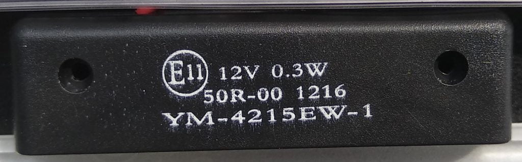 La imagen muestra una etiqueta impresa en un componente electrónico de color negro, específicamente el modelo YM-4215EW-1. En el centro de la etiqueta se detallan las especificaciones técnicas del dispositivo: 12V de voltaje, 0.3W de potencia, y el código de homologación E11, que indica cumplimiento con normativas europeas. También se visualiza el número de referencia 50R-00 1216, que puede corresponder al número de serie o lote de fabricación.Este tipo de componentes suele estar integrado en sistemas eléctricos o automotrices, y la información visible es crucial para tareas de mantenimiento, reparación, identificación o reemplazo de piezas.